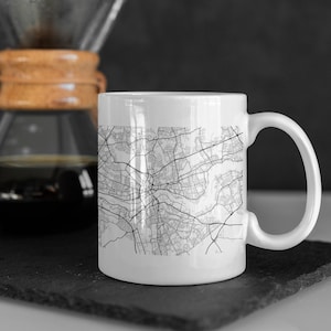 May include: White ceramic mug with a black and white city map design. The mug has a handle and is sitting on a black slate surface.