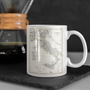 以下が含まれることがあります： 白磁のマグカップに、ヴィンテージのイタリア地図がプリントされています。地図には、国の海岸線、主要都市、地理的特徴が示されています。マグカップには、右側に持ち手があります。