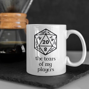 Peut inclure: Une tasse en céramique blanche avec une illustration en noir et blanc d'un dé à vingt faces et le texte "the tears of my players".