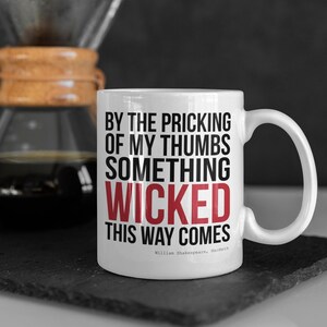 Puede incluir: Taza de cerámica blanca con una cita de Macbeth de Shakespeare en negro y rojo: "By the pricking of my thumbs, something wicked this way comes."