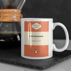 Op de afbeelding: Witte keramische mok met een ontwerp dat lijkt op een Penguin Books paperback. De omslag is oranje en heeft de tekst "I AM READING PLEASE GO AWAY" en een pinguïnlogo.