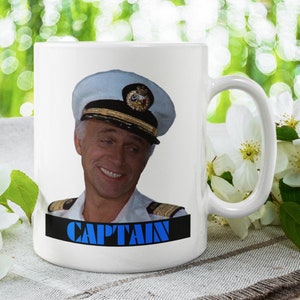 Könnte beinhalten: Weiße Keramik-Kaffeetasse mit einem Farbbild eines Mannes in einem Kapitänshut und Uniform. Der Text "CAPTAIN" ist in blauen Buchstaben auf einem schwarzen Balken unter dem Bild gedruckt.