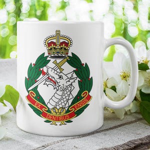 May include: White ceramic mug featuring a detailed emblem with a dragon, crown, and sword design. The emblem is surrounded by green laurel leaves and red banners with text. The mug has a white handle.