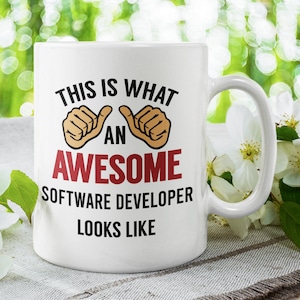 Może przedstawiać: Biały kubek ceramiczny z czarno-czerwonym wzorem, na którym widnieje napis "Tak wygląda niesamowity programista". Kubek ma dwa symbole kciuków w górę po obu stronach słowa "an".