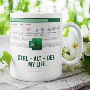 Peut inclure: Mug en céramique blanche avec un design de feuille de calcul Excel et le texte "CTRL + ALT + DEL MY LIFE" en vert. La tasse a une anse blanche. Des fleurs blanches sont en arrière-plan.