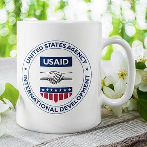 Könnte beinhalten: Eine weiße Keramiktasse mit einem blauen und roten Logo, auf dem "United States Agency for International Development" und "USAID" steht. Das Logo zeigt einen Händedruck zwischen zwei Händen.