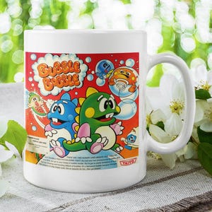 May include: White ceramic mug featuring a colourful graphic of the video game Bubble Bobble. The graphic depicts two cartoon characters, one green and one blue, blowing bubbles. The text "Bubble Bobble" is printed in red and white.
