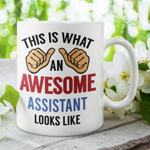 May include: White ceramic coffee mug with a blue, red and black design that says "This is what an awesome assistant looks like". The mug has two thumbs up symbols on either side of the word "an".
