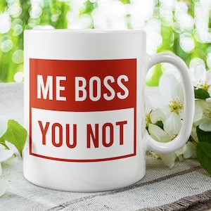 May include: White ceramic mug with a red square that says "ME BOSS YOU NOT" in white text. The mug is sitting on a white and grey textured surface with white flowers.