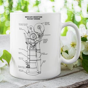 May include: White ceramic mug featuring a detailed black and white diagram of the Mercury-Redstone rocket engine. The mug has a large handle and is labeled with various engine components. The background includes white flowers and green foliage.