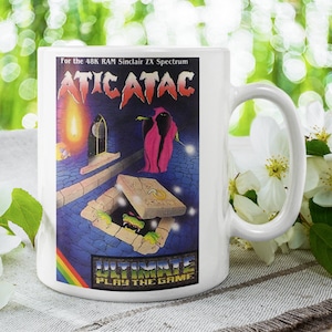 Könnte beinhalten: Weiße Keramiktasse mit einem bunten Bild eines Videospiels namens "Atic Atac" für den 48K RAM Sinclair ZX Spectrum. Das Bild zeigt eine dunkle Gestalt in einem roten Gewand, eine Falltür und eine grüne Kreatur.
