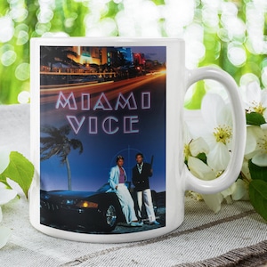 Può includere: Tazza in ceramica bianca con un'immagine colorata di una città notturna, due uomini in piedi accanto a un'auto nera e il testo "Miami Vice".