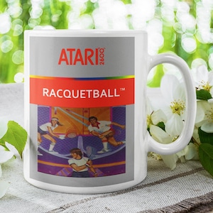 May include: White ceramic mug featuring the Atari 2600 logo and the game title "Racquetball." The mug's design includes a colorful graphic of the game's gameplay. The mug has a large handle and is set against a blurred background of flowers.