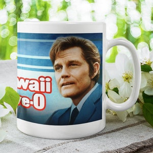 Puede incluir: Taza de cerámica blanca con un gráfico azul y blanco de un hombre con el texto "Hawaii Five-0" impreso en ella.