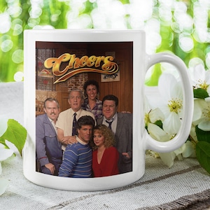 May include: White ceramic coffee mug with a colorful image of the cast of the TV show "Cheers" printed on the front. The image includes the show's title in gold lettering.