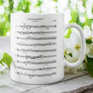 以下が含まれることがあります： 「Concerto from Op.85」の楽譜がプリントされた白いセラミックマグ。マグには大きなハンドルがあり、白い花と緑の葉に囲まれています。背景はぼやけています。