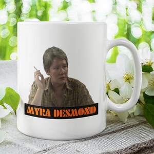 May include: White ceramic mug with a color image of a person with short brown hair holding a cigarette. The mug has the text "MYRA DESMOND" printed in orange letters on a black background.