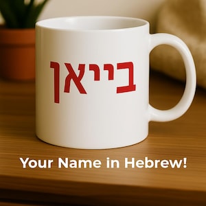 Puede incluir: Taza de cerámica blanca con una inscripción hebrea roja. La taza tiene un asa grande y está sobre una superficie de madera. El texto "Your Name in Hebrew!" está impreso en la parte inferior de la imagen.