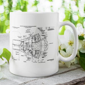 May include: White ceramic mug featuring a detailed black and white diagram of the Mercury spacecraft interior arrangement. The mug has a large handle and is set against a blurred green background with white flowers.