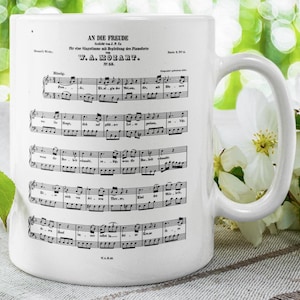 May include: White ceramic mug featuring a black and white image of sheet music with the title "An die Freude" by W.A. Mozart. The music is for a single voice with piano accompaniment.