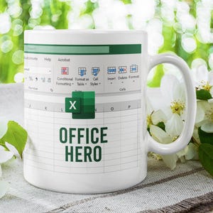 May include: White ceramic mug with an Excel spreadsheet design and the words "OFFICE HERO" in green. The mug has a white handle. White flowers and greenery are in the background.