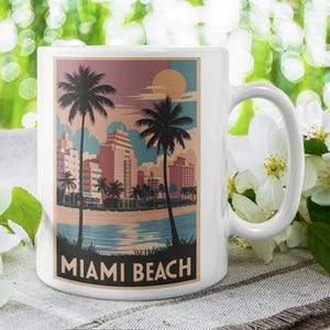 May include: White ceramic mug featuring a Miami Beach design. The design includes palm trees, buildings, and the text "MIAMI BEACH" in a black rectangle. The background has a sunset color scheme.