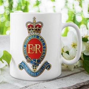 May include: White ceramic mug featuring the Royal Horse Artillery emblem. The emblem includes a crown, the initials "ER", and the motto "Honi Soit Qui Mal Y Pense". The mug has a curved handle and is set against a blurred green background.