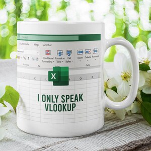 May include: White ceramic mug with an Excel spreadsheet design and the text "I ONLY SPEAK VLOOKUP" in green. The mug has a white handle. The image includes white flowers and green foliage.