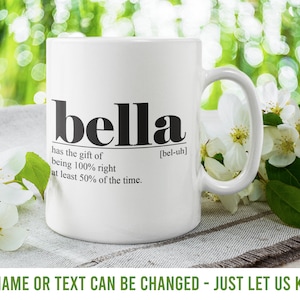 Könnte beinhalten: Weiße Keramiktasse mit schwarzem Text, der "bella" lautet und eine Definition des Namens. Die Tasse steht auf einer hellbraunen Oberfläche mit weißen Blumen.