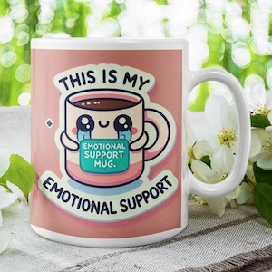 May include: White ceramic mug with a pink background and a cartoon illustration of a mug with a face and the text "This is my emotional support mug. Emotional Support".