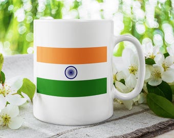 Op de afbeelding: Witte keramische mok met het Indiase vlagontwerp. De mok heeft een handvat en is versierd met oranje, witte en groene strepen en een blauw wielembleem. De mok staat op een getextureerd oppervlak met witte bloemen.