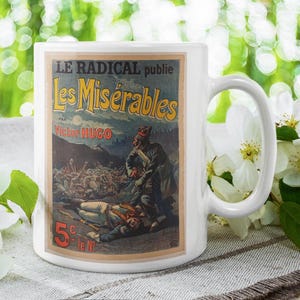 May include: White ceramic mug featuring a vintage poster design for Victor Hugo's "Les Misérables." The poster art depicts a scene with soldiers and the text "LE RADICAL publie." The mug has a curved handle.