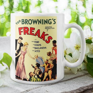 May include: White ceramic mug displaying the movie poster for Tod Browning's "Freaks." The poster art features illustrations of characters and the film's title in red and yellow. The mug is set against a blurred green background with white flowers.