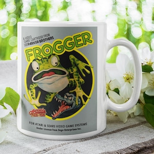 May include: White ceramic mug featuring the Frogger video game logo. The mug has a large handle and displays the game's frog character in a yellow circle. Text includes "Frogger" and "A Video Game Cartridge From Parker Brothers."