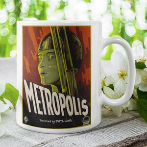May include: White ceramic mug with a vintage "Metropolis" film poster design. The poster art features a woman's profile, the film's title, and the director's name. The mug is set against a blurred green backdrop, with white flowers in the foreground.