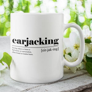 May include: White ceramic mug with a black graphic that defines "earjacking" as forcing others to listen to bad music or conversation, often found on London buses. The mug is sitting on a white surface with white flowers.
