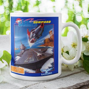 Puede incluir: Taza de cerámica blanca con una imagen colorida de un videojuego llamado "Carrier Command" que presenta una aeronave futurista volando sobre un acorazado. La taza tiene un borde azul y blanco y el texto "Rainbird" en la parte inferior.