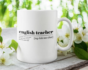 May include: White ceramic mug with black text that reads "english teacher" and a definition of the word. The mug is sitting on a white linen surface with white flowers.