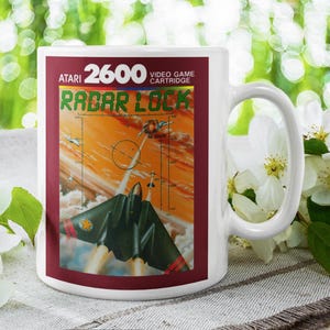 Puede incluir: Taza de cerámica blanca con el cartucho de videojuego Atari 2600 "Radar Lock". El diseño de la taza incluye el título del juego, un gráfico de un avión y un fondo naranja y amarillo vibrante. La taza está sobre un fondo verde borroso.