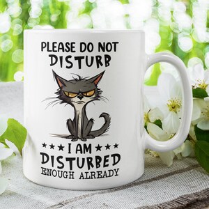 Könnte beinhalten: Eine weiße Keramik-Kaffeetasse mit einer Illustration einer schwarzen und grauen Comic-Katze. Die Tasse trägt den Text "Bitte nicht stören. Ich bin schon genug gestört."