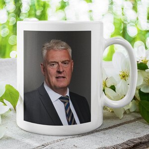 May include: White ceramic mug featuring a photograph of a man in a suit and tie. The man is looking at the camera and has short, dark hair. The mug has a handle on the right side.