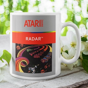 May include: White ceramic mug featuring the Atari 2600 game "Radar" with a colorful graphic design. The mug has a white handle and is set against a blurred green background with white flowers.
