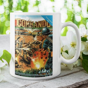 Könnte beinhalten: Weiße Keramiktasse mit einem bunten Bild eines Videospielcharakters, der eine Waffe hält. Das Bild stammt aus dem Spiel Commando für den Commodore 64. Der Text "Commodore 64" und "Elite" ist auf der Tasse sichtbar.