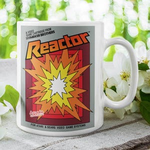 May include: White ceramic mug featuring the "Reactor" video game cartridge design. The design includes a burst of orange and yellow colors, with the game title and branding. The mug is for Atari & Sears video game systems.