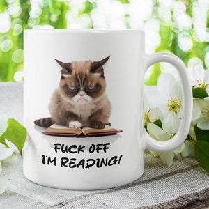 May include: White ceramic coffee mug with a grumpy cat illustration reading a book. The text on the mug reads "F**k Off I'm Reading!"