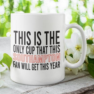 Può includere: Tazza in ceramica bianca con testo nero che recita "This is the only cup that this Southampton fan will get this year". La tazza è appoggiata su una superficie testurizzata bianca e grigia con fiori bianchi.