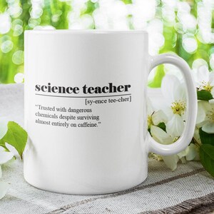 May include: White ceramic mug with the words "science teacher" and a definition printed in black. The mug also includes the quote: "Trusted with dangerous chemicals despite surviving almost entirely on caffeine." The mug is next to white flowers.
