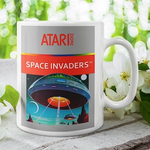Könnte beinhalten: Weiße Keramik-Tasse mit dem Atari 2600 Logo und Space Invaders-Design. Die Tasse zeigt eine farbenfrohe Illustration eines Raumschiffs und den Titel des Spiels. Die Tasse hat einen großen Henkel und steht vor einem verschwommenen floralen Hintergrund.