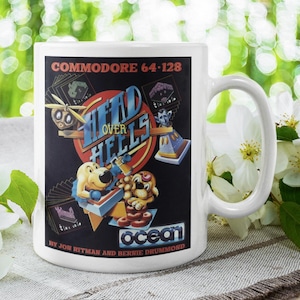 Puede incluir: Taza de cerámica blanca con una imagen en blanco y negro de un cartucho de videojuego para Commodore 64-128. El juego se llama "Head Over Heels" y es de Jon Ritman y Bernie Drummond. La imagen también incluye el logotipo de Ocean Software.