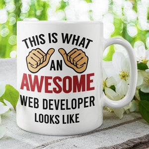 May include: White ceramic coffee mug with a black and red design. The mug has the text "This is what an awesome web developer looks like" with two thumbs up emojis.